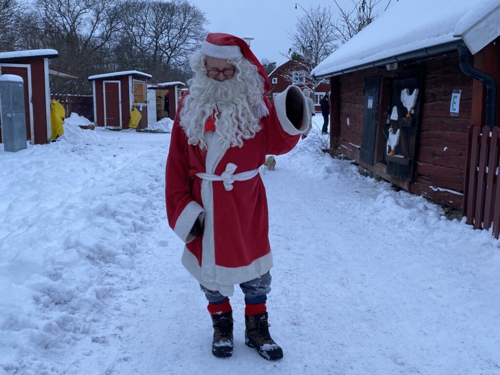 Tomte står på en snöig väg och pekar, röd lada till höger om honom